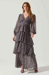 A woman is wearing a long, flowing dress made of sheer fabric with a small floral pattern in purple, white, and black. The dress features a V-neckline, long puffed sleeves with elastic cuffs, and a tiered, ruffled skirt that reaches the floor.